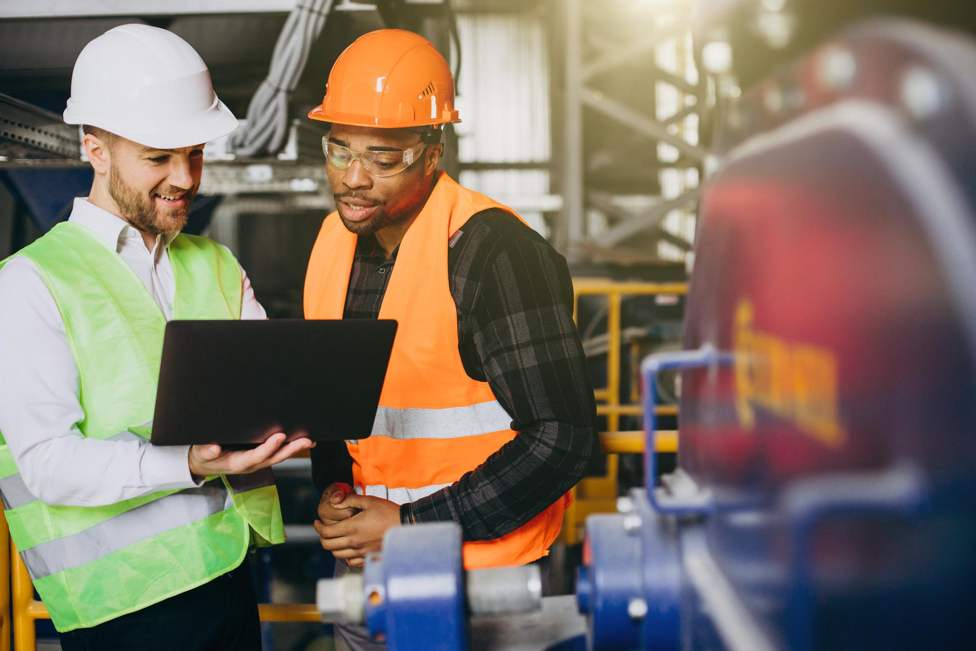 Two factory engineers wearing safety vests and hard hats review production data on a laptop while standing beside industrial equipment in a modern manufacturing facility.